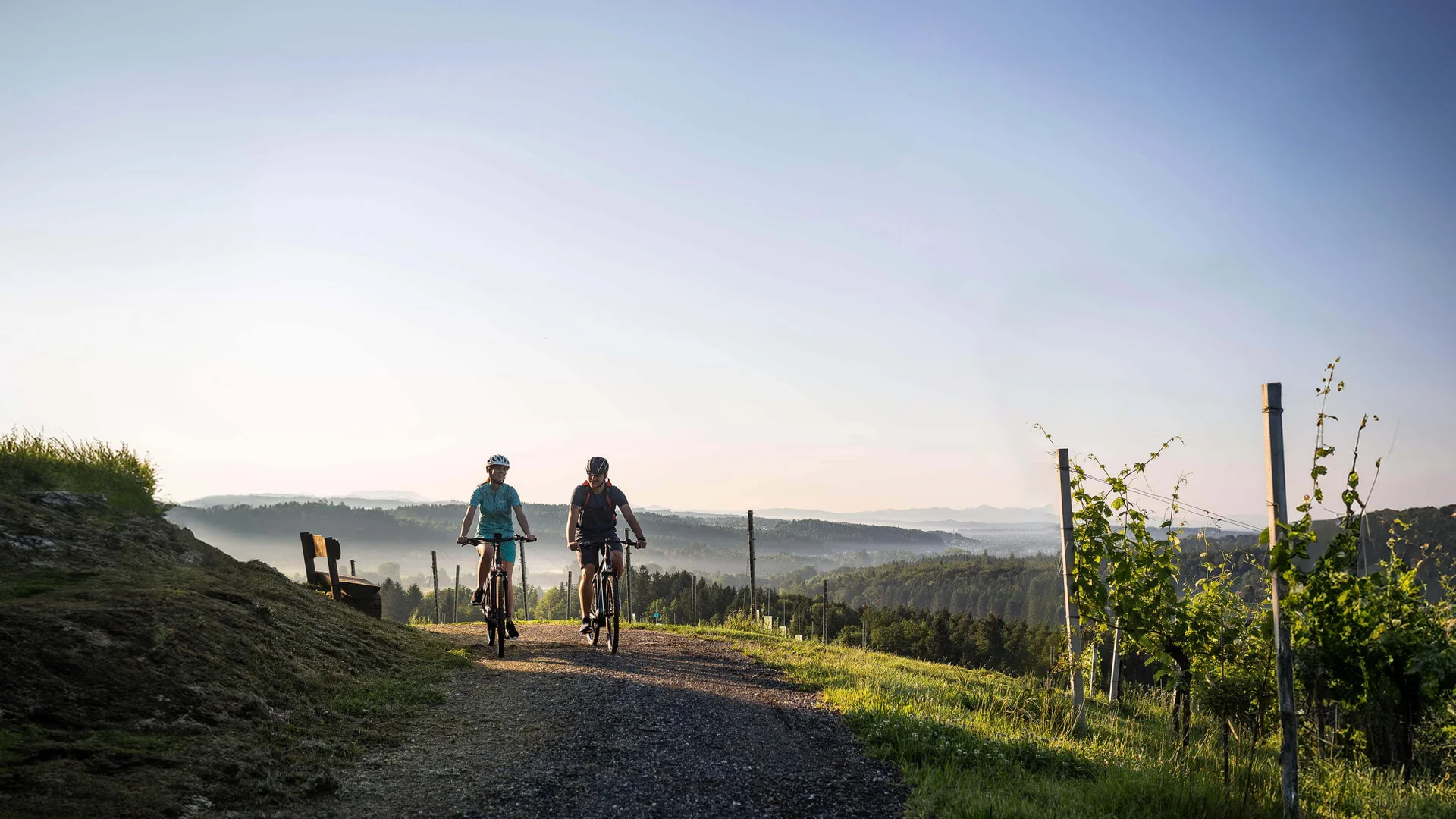 Velontour Zwei Radfahrer fahren auf einem Weg durch eine hügelige Landschaft bei klarem Himmel