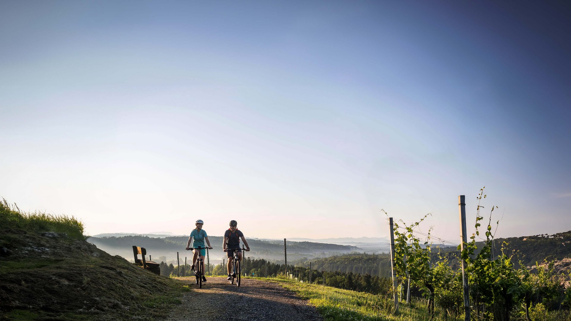 Velontour Zwei Radfahrer fahren auf einem Weg durch eine hügelige Landschaft bei klarem Himmel