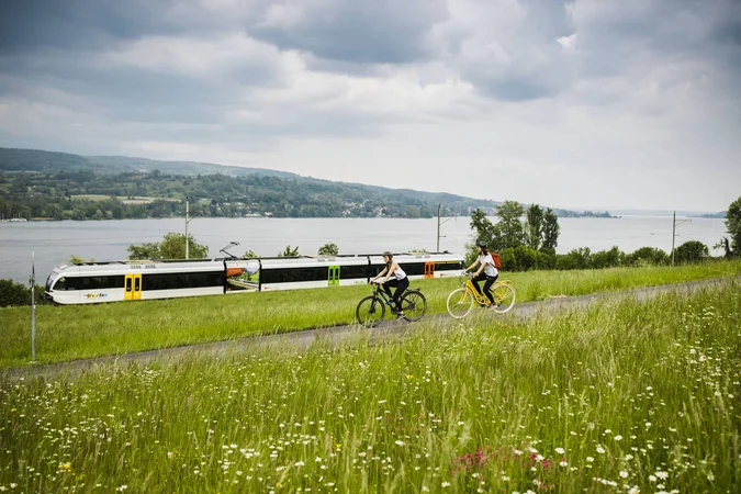 Velontour © Ivo Scholz Zwei Radfahrer fahren auf einem Weg neben einer Bahnstrecke am See