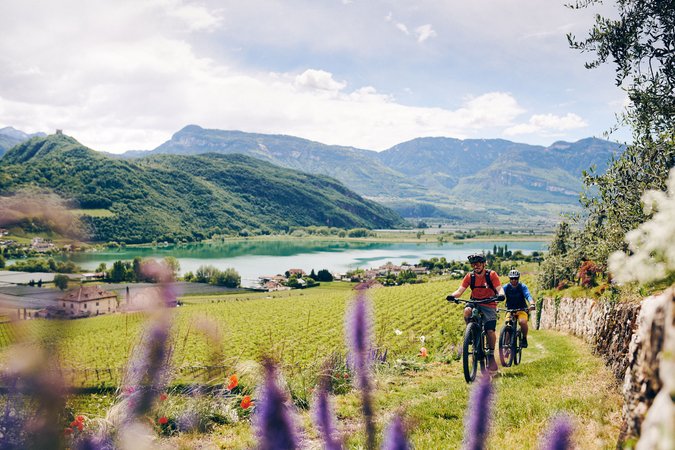 Velontour © IDM Südtirol - Manuel Ferrigato Zwei Radfahrer fahren durch eine grüne Landschaft mit Bergen und See im Hintergrund