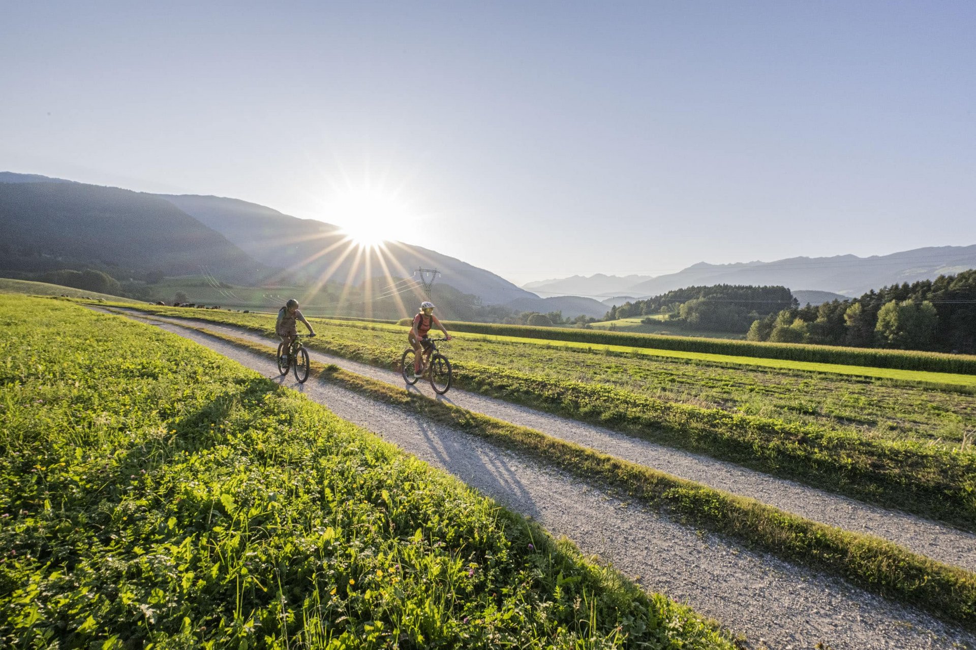 Velontour © Harald Wisthaler Zwei Radfahrer fahren auf einem Feldweg mit Bergen und Sonnenuntergang im Hintergrund