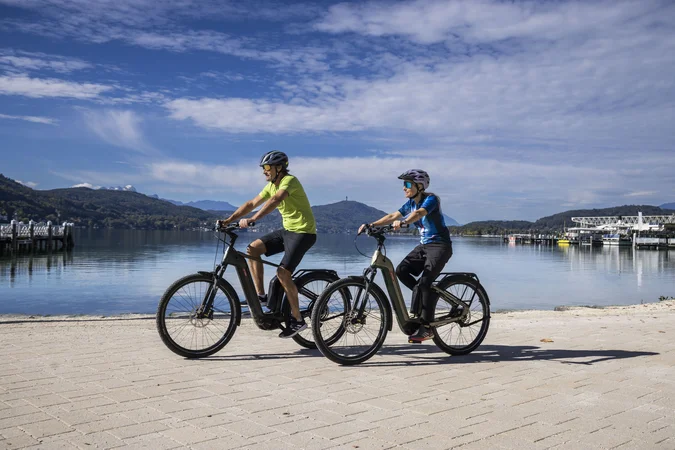 Velontour © Velontour - Heiko Mandl Zwei Personen fahren mit E-Bikes an einem See mit Bergen im Hintergrund