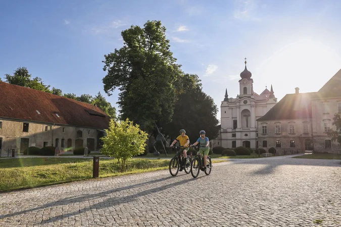 Velontour © Velontour Zwei Radfahrer bei sonnigem Wetter vor historischen Gebäuden und Bäumen
