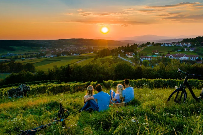 Velontour © TV Thermen & Vulkanland - Mias Photoart Vier Personen sitzen auf einer Wiese und schauen auf den Sonnenuntergang über dem Dorf