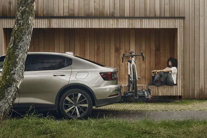 Velontour © Thule Person sitzt neben einem Fahrrad an einer Holzfassade, daneben steht ein Auto