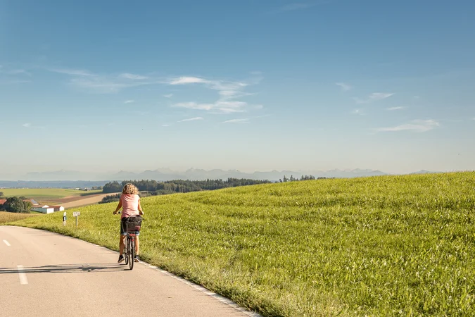 Velontour © Tourismusverein Inn-Salzach Frau fährt Fahrrad auf Landstraße neben grünem Hügel bei klarem Himmel