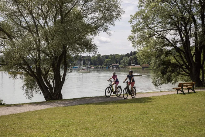 Velontour © Velontour Zwei Radfahrer auf Weg am Seeufer mit Booten und Bänken unter Bäumen