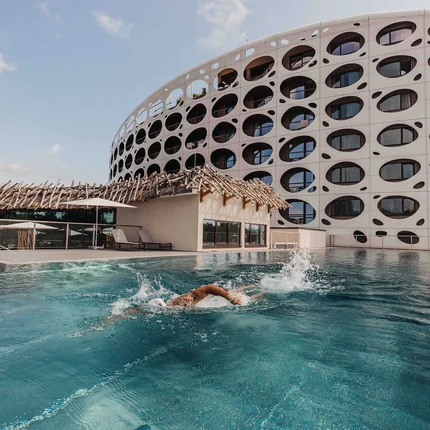 Das Seepark Wörthersee Resort © Daniel Waschnig Person schwimmt im Außenpool vor einem modernen Gebäude mit runden Fenstern