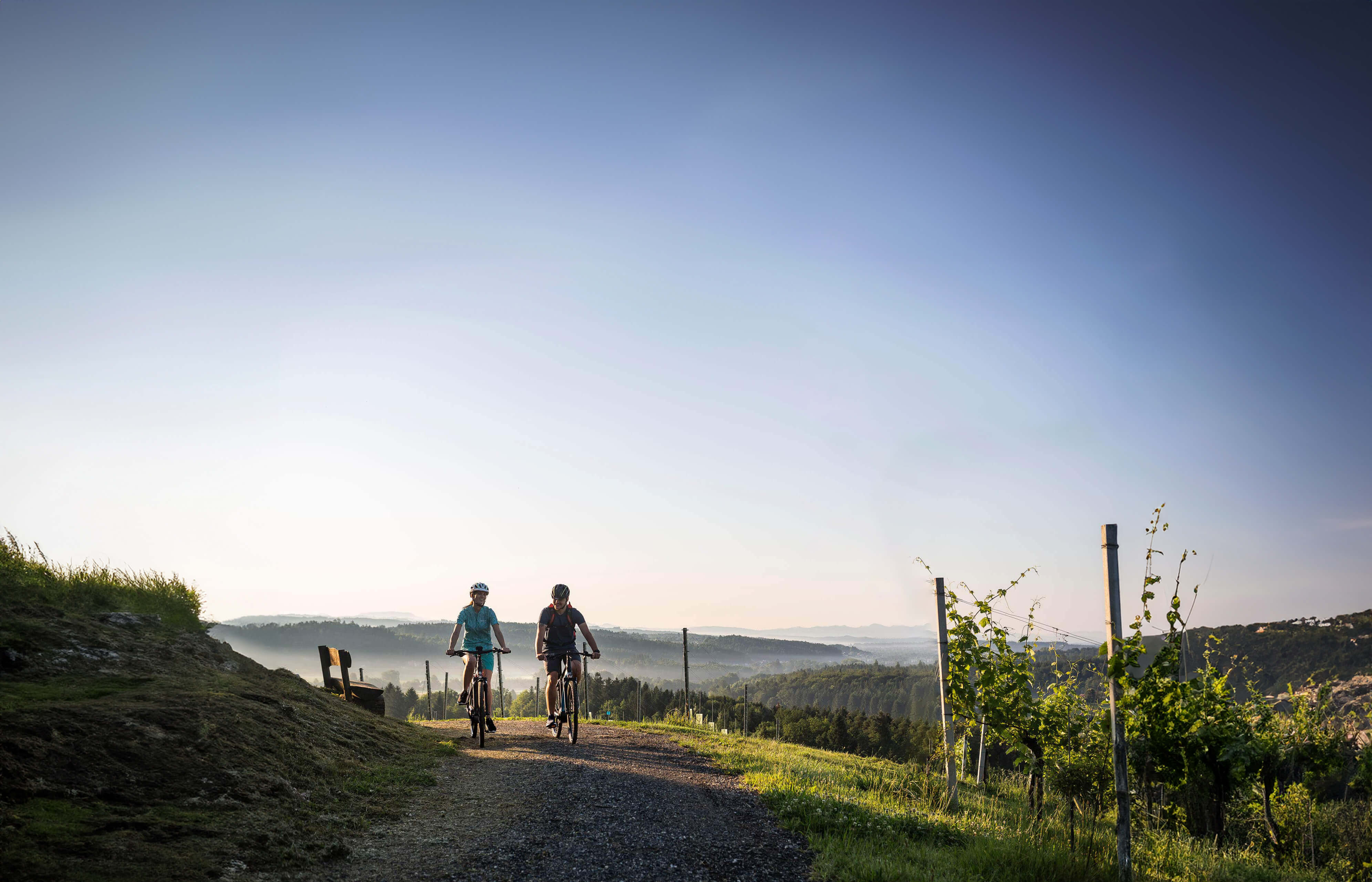 Velontour Zwei Radfahrer fahren auf einem Weg durch eine hügelige Landschaft bei klarem Himmel